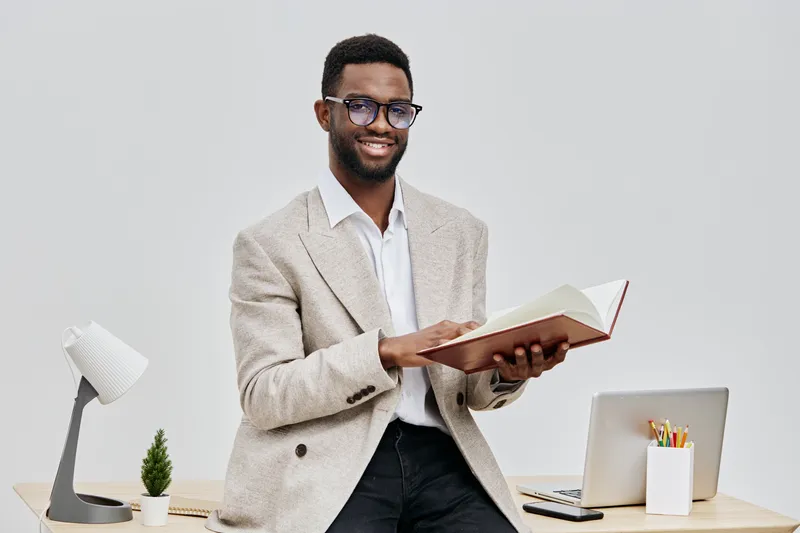 Professional smiling young African-American man in beige blazer holding open book in office environment with desk, laptop, and stationery, conveying learning and education concept.