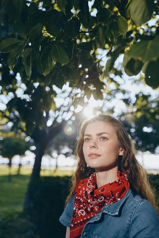 young woman standing outdoors under green leaves with sunlight filtering through, wearing a red scarf and denim jacket, calm and thoughtful expression