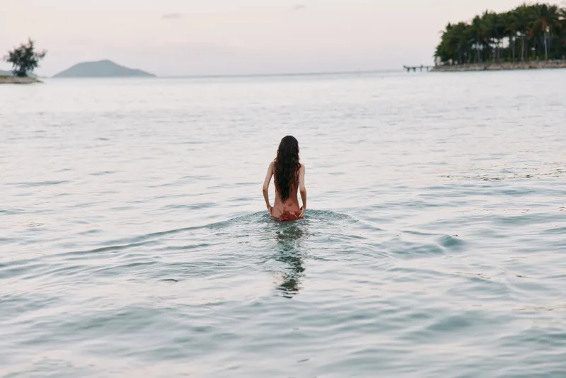 A solitary figure stands waist deep in tranquil sea, back turned, facing distant horizon as soft light brushes the water and coastline, creating serene marine solitude.