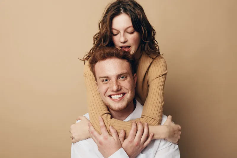 Smiling redhead man with freckles embraces happy woman with curly hair in beige sweater. Couple shows affection and warmth on neutral background. Concept of love, friendship, and togetherness.