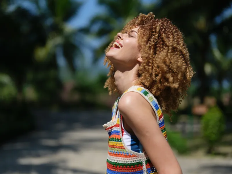 happy young woman with curly hair laughing outdoors, wearing colorful striped top, enjoying sunny day with blurred tropical trees in background, carefree summer moment