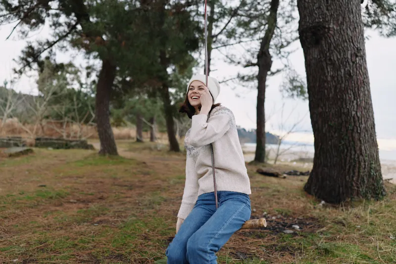 Woman outdoors in a forest by the coast, wearing a cozy sweater and jeans, smiling softly while enjoying nature and a relaxed pose.