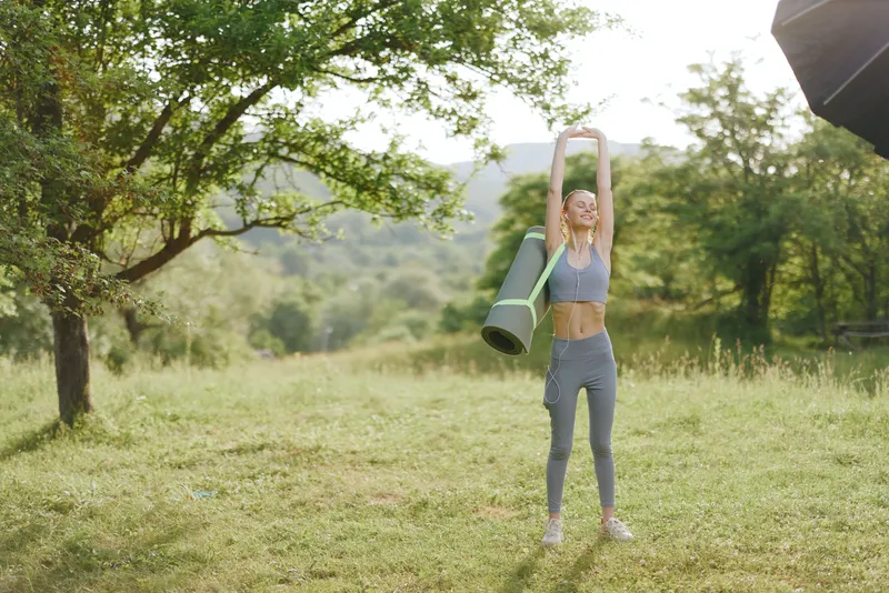 Young woman practicing yoga outdoors in athletic wear, stretching with a yoga mat in a serene nature setting, embodying health and wellness