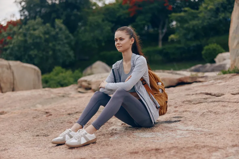 Young woman in athletic wear sitting on rocks, enjoying nature with a backpack, showcasing fitness and relaxation in a serene outdoor setting