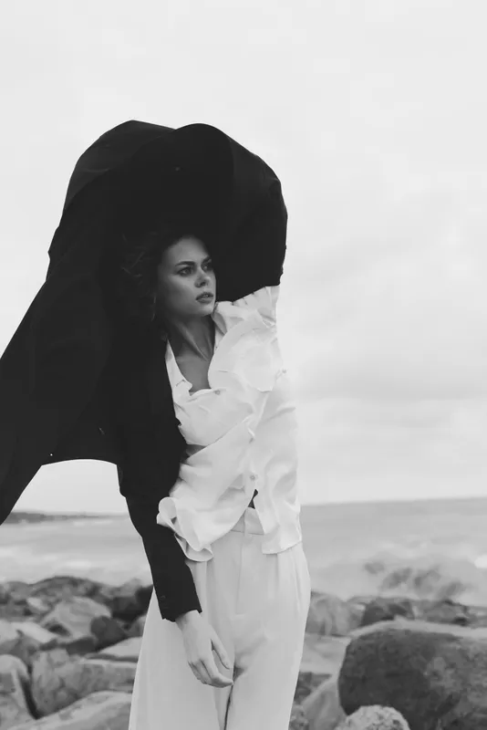 Wind swept fashion on a coastal backdrop, a model in a white dress and wide hat, captured in monochrome editorial mood among rocks by sea