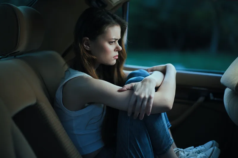 Pensive young woman sitting in a car, reflecting with a thoughtful expression, wearing casual clothing, atmospheric evening mood.