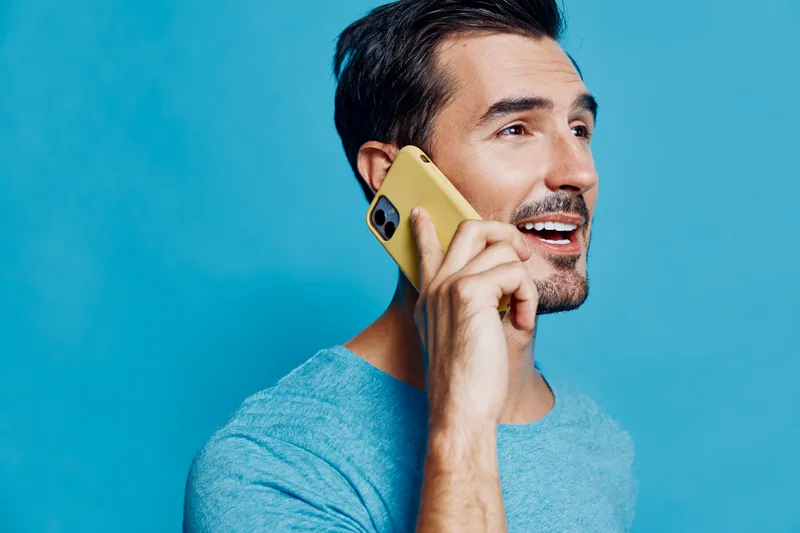 Smiling man talking on the phone while wearing a blue t-shirt against a vibrant blue background, expressing joy and connection in a modern lifestyle.