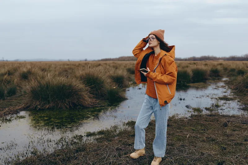 Woman lifestyle outdoors in rural field path, wearing orange jacket, knit hat and sunglasses, walking with relaxed smile, hands in pockets, cloudy daylight, full body, natural scene