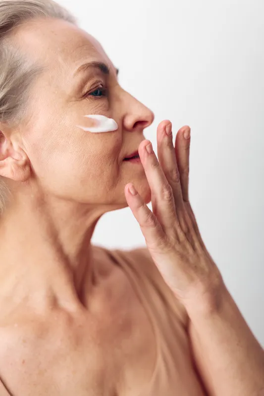 Mature woman applies white face cream on cheek, showing skincare routine and self-care. Soft natural light highlights healthy skin and beauty in minimalist setting.