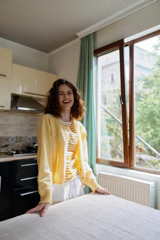 A smiling woman stands in a sunlit kitchen, wearing a yellow cardigan over a striped top, resting hands on a table. A candid lifestyle scene conveying warmth, home comfort, and everyday life. The