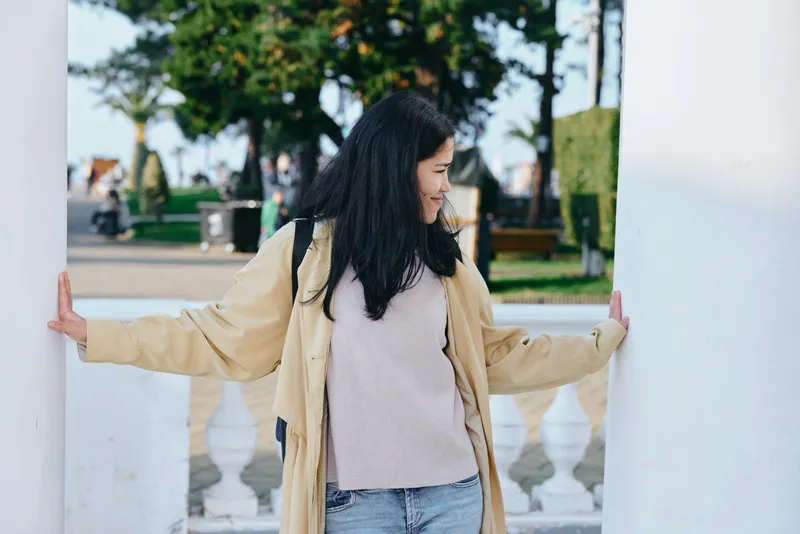 Woman stands between columns in an outdoor park, wearing a beige coat and pink sweater with jeans, capturing a relaxed street style scene under bright daylight and calm ambience in a sunny urban