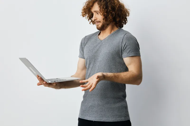 Young man with curly hair in casual gray t shirt, expressing confusion while using a laptop against a minimalist backdrop, ideal for technology themes