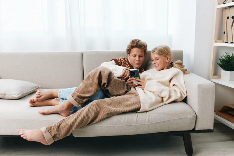 Two friends lounge on a light sofa, sharing a phone and relaxed smiles, capturing casual home leisure and digital connection in a bright, cozy living room.
