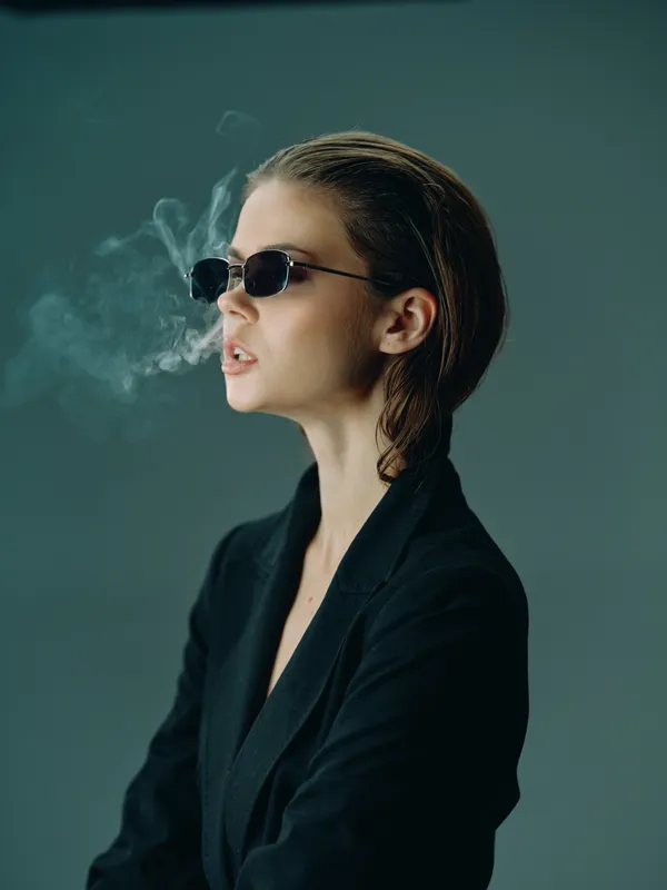 Young stylish woman wearing sunglasses smoking with smoke cloud on moody dark background