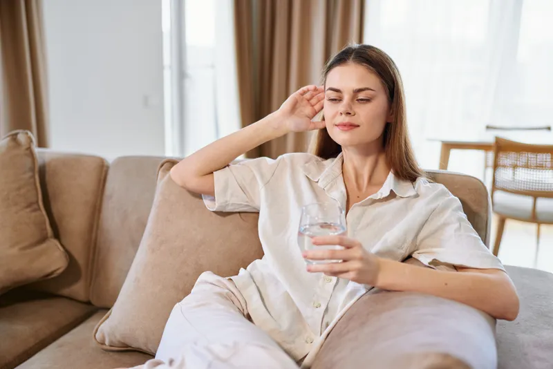 Woman on sofa holding a glass of water, relaxation at home in a bright living room, casual clothing and peaceful expression, hydration and self care during a quiet afternoon.
