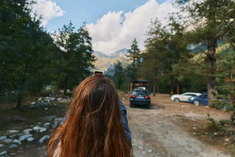 A person with long hair stands in a forested roadside area, capturing a photo with a handheld device. The scene includes trees, parked cars, and distant mountains, conveying adventure, exploration,