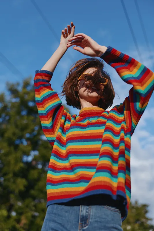 Young woman in a rainbow sweater and sunglasses outdoors, arms raised under blue sky. Early 20s model smiling in a casual outfit with short brown hair, urban park lifestyle fashion portrait.