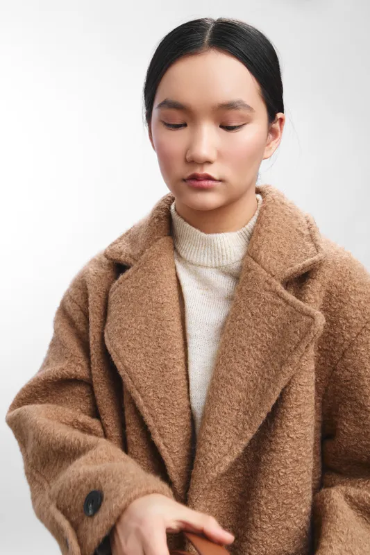Asian woman in a warm beige coat stands in a studio, looking down with a calm, thoughtful expression. The minimalist portrait highlights texture, subtle fashion, and quiet confidence.