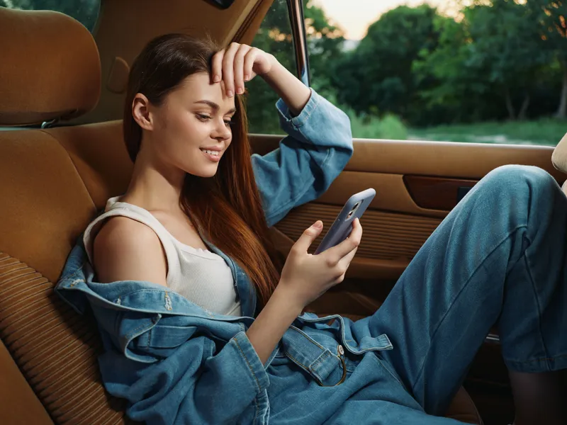 Young woman in denim outfit smiling while using smartphone in car, surrounded by greenery, conveying a relaxed and casual mood.