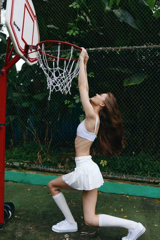 A young woman in a white sports outfit performs a slam dunk at a basketball court, showcasing athleticism and energy against a lush green background