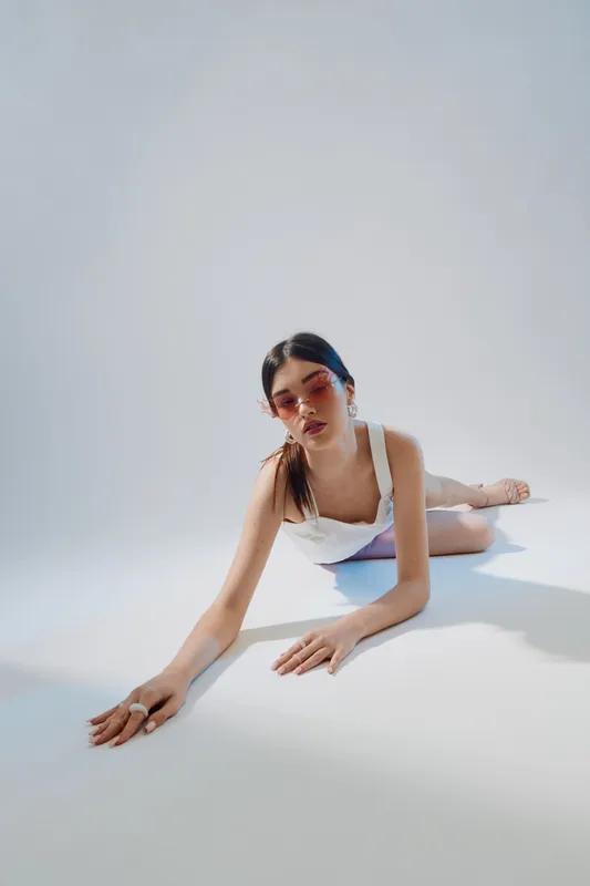 Young woman in white fashion outfit lies on the floor wearing pink sunglasses. Minimalist studio setting with soft lighting highlights beauty and style concepts.