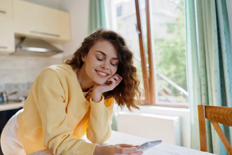 woman lifestyle home smiling cozy interior sunlight device casual moment. A relaxed woman indoors wearing a yellow sweater, enjoying a bright window seat, creating a warm, intimate lifestyle moment.