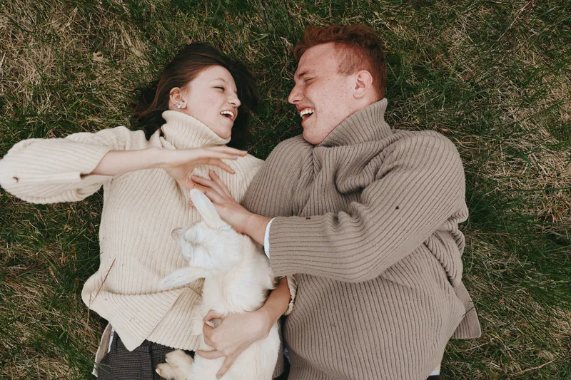 Cheerful couple outdoors, lying on grass, laughter and warm sweaters, sunlit moment, relaxed mood, affectionate interaction, peaceful park scene, pet dog nearby soft, natural colors, lush greenery