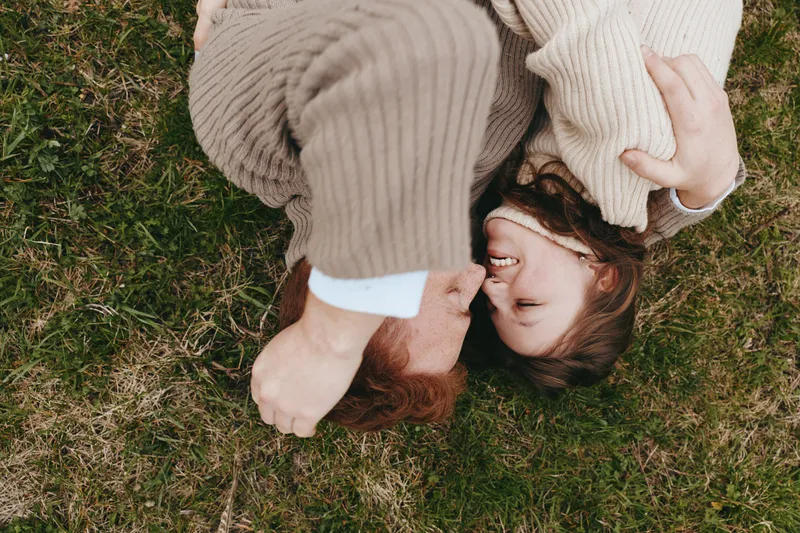 Couple lying on grass wearing cozy sweaters, smiling and embracing in a warm outdoor moment, natural light highlighting affectionate interaction and relaxed lifestyle.