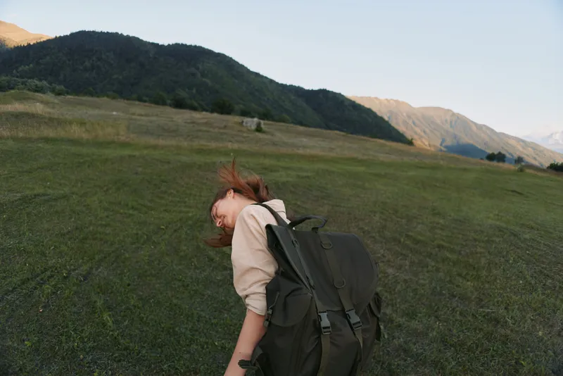 A young woman with a large backpack bends in a grassy field with distant mountains behind, capturing a moment of exploration and solitary travel.