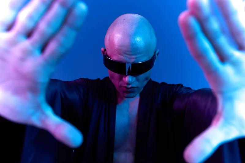 A bald man wearing sunglasses poses dramatically against a blue gradient background. The image captures a powerful and modern vibe, embracing tech-inspired aesthetics.
