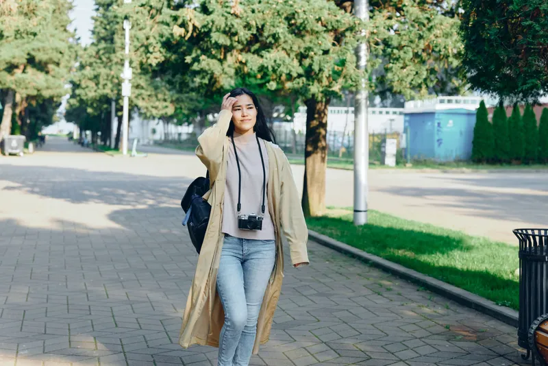 Woman walking along a sidewalk outdoors in a long coat, camera hanging from the neck, exuding casual fashion and travel vibes in an urban street scene today, vivid and candid
