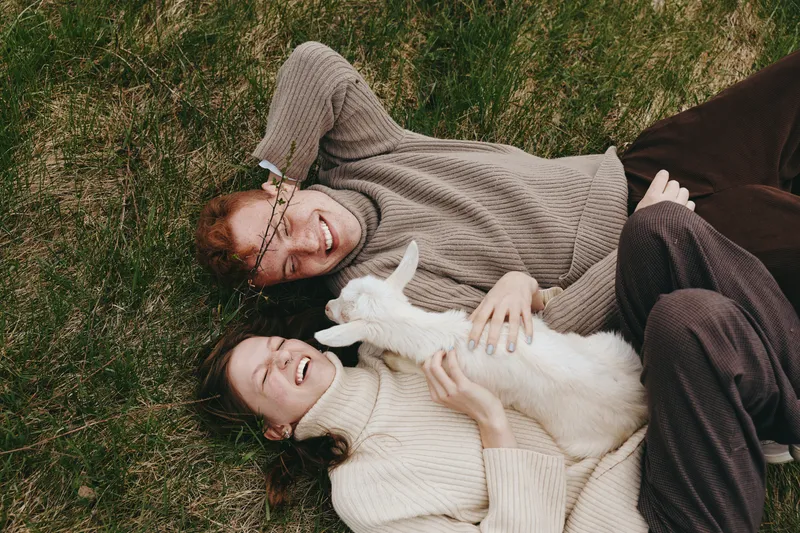 Cheerful couple outdoors, lying on grass with a playful white goat kid, smiling and cuddling, soft knit sweaters, warm sun, natural colors, relaxed lifestyle moment in nature