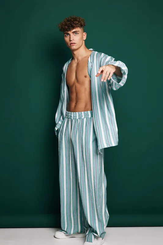 Panelitudinarian young man with curly hair wearing striped pajamas open shirt pointing at camera posing against green background with confident expression and relaxed stance.