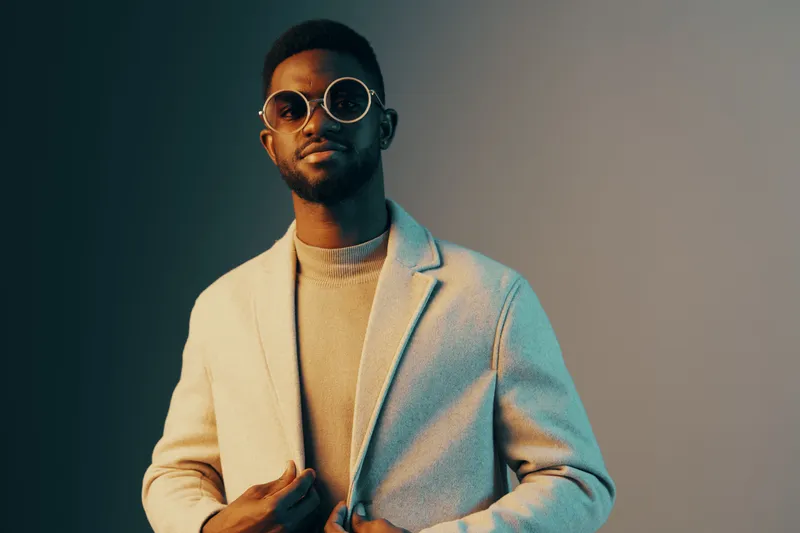 A stylish Black man in a trendy coat and sunglasses poses confidently against a gradient background, showcasing modern fashion and self assuredness
