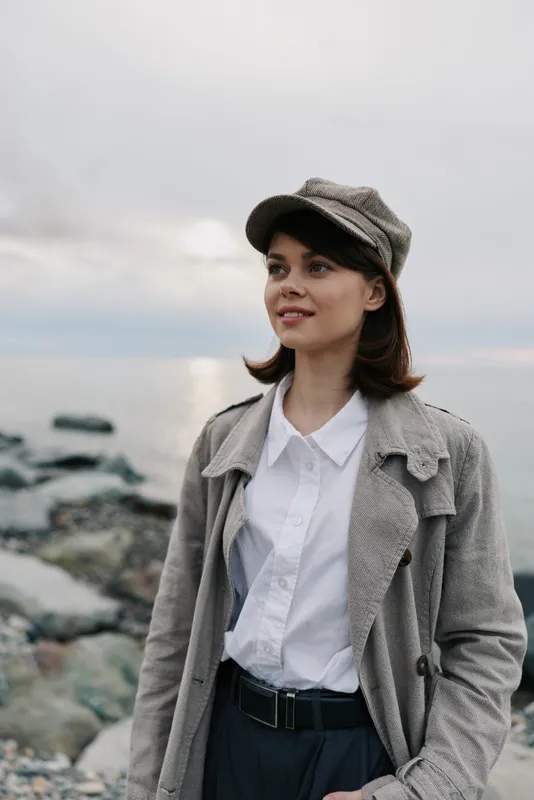 Young woman wearing a hat and coat stands near rocky shore looking to the side. Overcast sky and calm sea create a peaceful atmosphere. Concept of autumn fashion and nature.