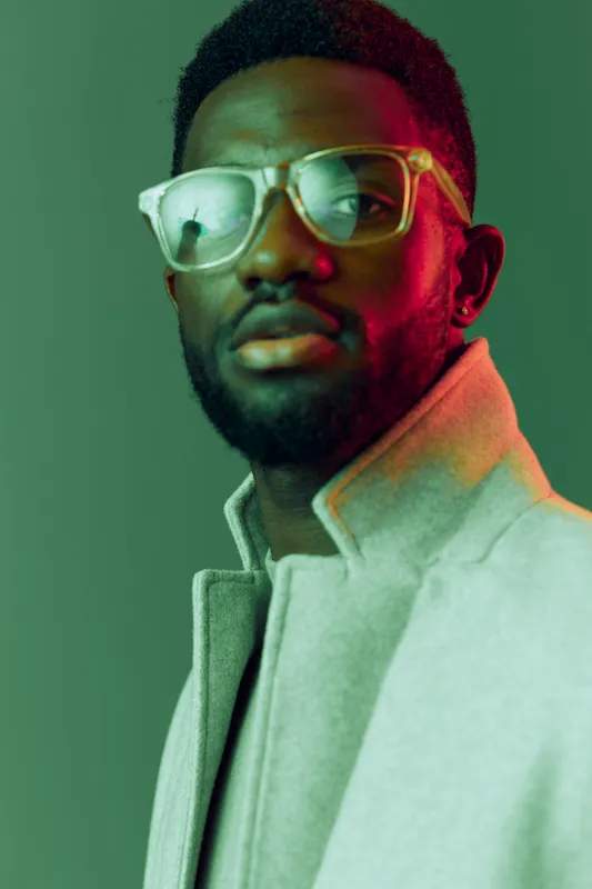 A stylish male model with glasses, wearing a fashionable coat against a vibrant green background, exuding confidence and elegance