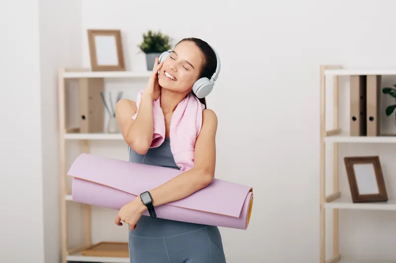 Fitness portrait of a smiling woman in athletic wear with headphones, hugging a pink yoga mat and towel in a bright room with shelves and plants, ready for workout.