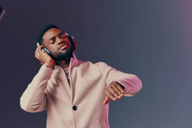 Young African man enjoying music with headphones in stylish coat, relaxed expression, against a gradient background, perfect for music and lifestyle themes
