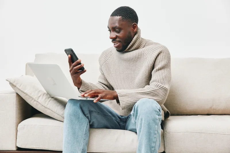 Young African man in cozy sweater using laptop while texting on smartphone, sitting on beige couch in modern home office environment