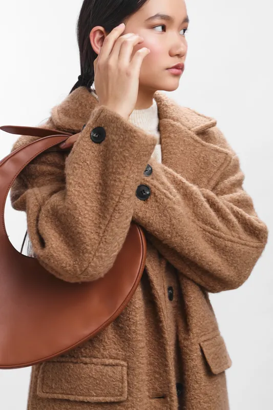 Asian woman wearing a brown wool coat and a curved leather bag stands in a clean studio, adjusting her hair and looking aside with calm, poised confidence.