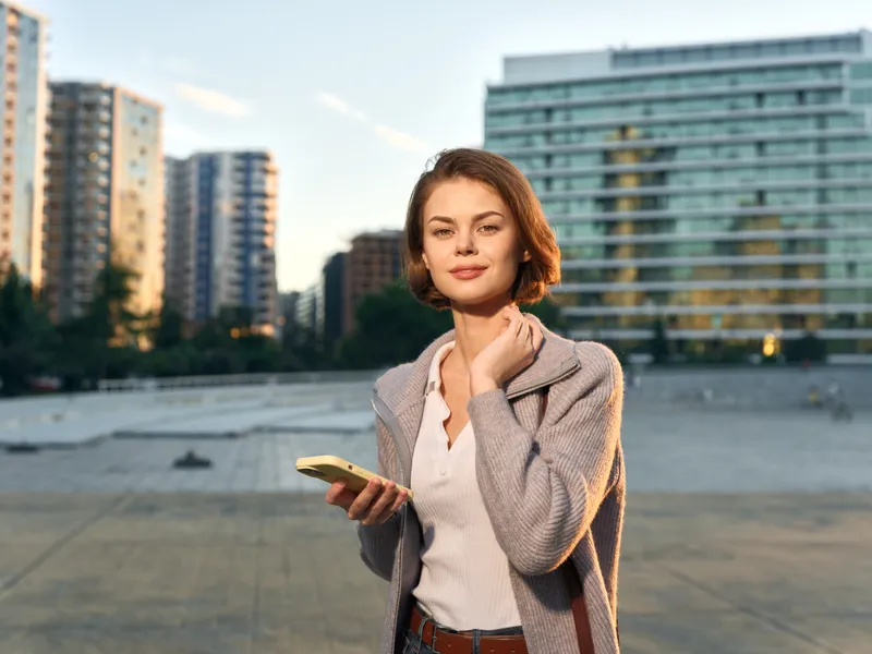 A cheerful professional woman stands outdoors in a modern cityscape, holding a smartphone. She smiles confidently, presenting a positive, connected, and dynamic work vibe.