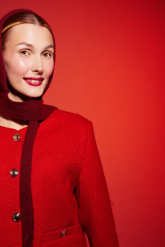 Smiling woman in red coat and dark scarf poses against red background. Studio portrait with warm lighting highlights her clear skin and confident expression. Concept of fashion and elegance.