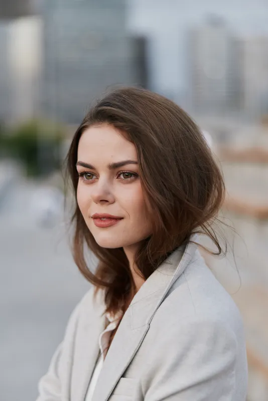 Professional woman in a light gray blazer with natural makeup, exuding confidence against an urban city background, embodying modern business style and ambition.