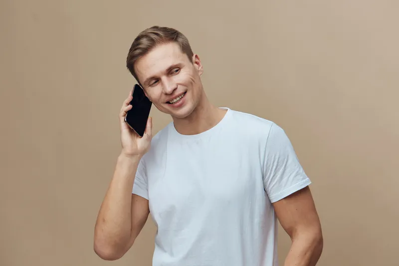 smiling young caucasian man wearing white t-shirt talking on phone isolated on beige background digital nomad concept technology communication lifestyle