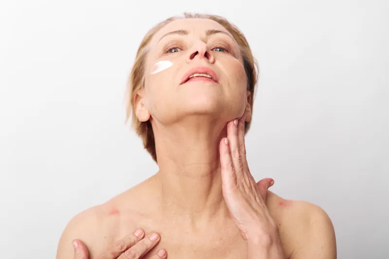 Mature woman applies cream on neck and face with visible skin care product on cheek. Clean white background highlights natural beauty and self care routine for aging skin and wellness concept.