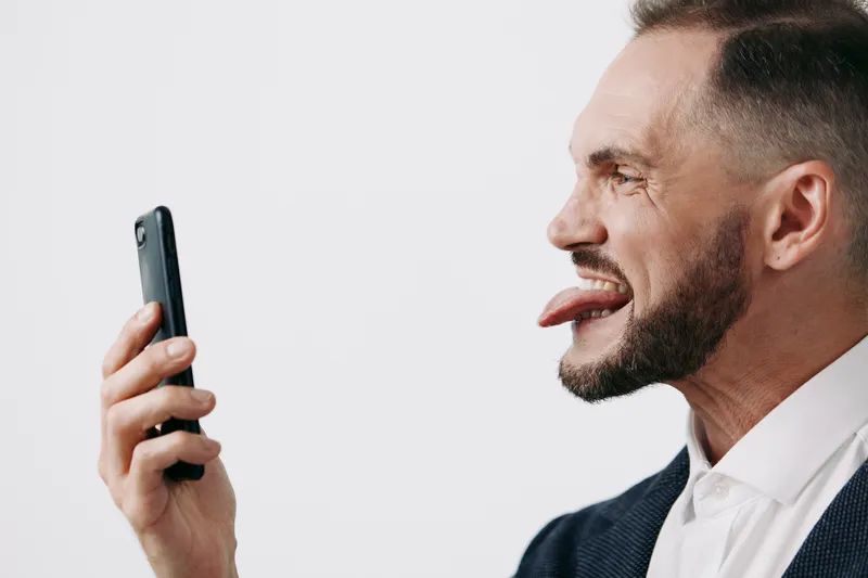 Professional business man engages with a smartphone on an isolated, clean background, showing playful humor and a focused, expressive mood during a digital interaction.