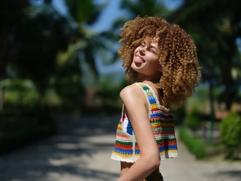 cheerful young woman with curly hair wearing colorful striped top smiling and enjoying sunny day outdoors, tropical palm trees in background, summer lifestyle portrait