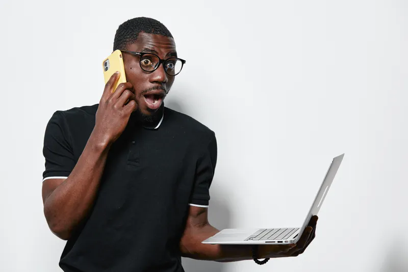 African American man with dark skin and glasses holding laptop and smartphone looking surprised in casual black shirt against white background. Technology, communication and expression.