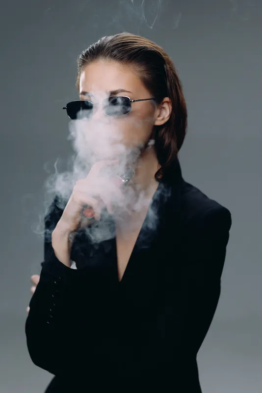 Woman in sunglasses blowing smoke with stylish black outfit and confident look on gray background