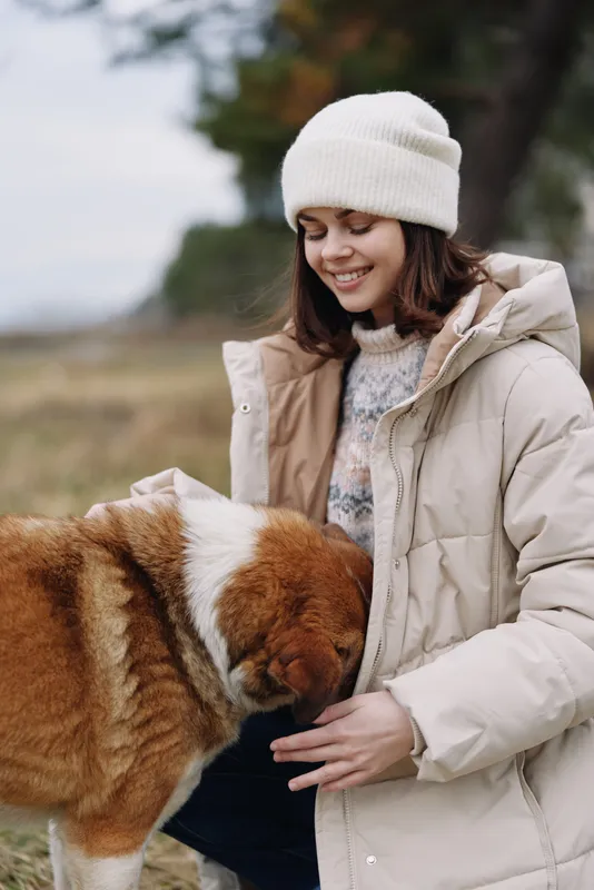 Woman in a warm coat outdoors pets a large dog on a chilly day, sharing a moment of companionship and care amid open fields, soft sunlight, and a peaceful park setting with a gentle breeze